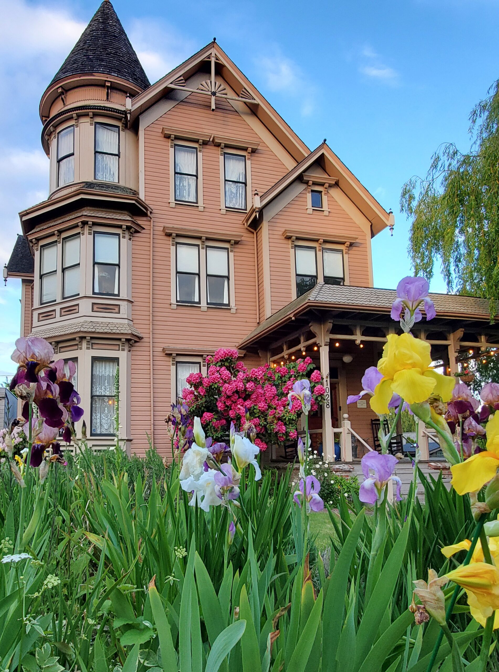 Exterior of the Adams Pragge House, a three-story Victorian with turret, pictured in May with abundant flowers bloomingforeground.
